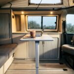 Blick in einen Campervan bei Regen, auf dem Tisch ein Laptop und eine Tasse heiße Schokolade, der Platz davor ist leer