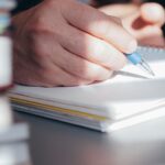 Best Possible Self Übung: Eine Hand schreibt im hellen Tageslicht mit Kugelschreiber in ein Notizbuch, daneben ein Stapel Bücher